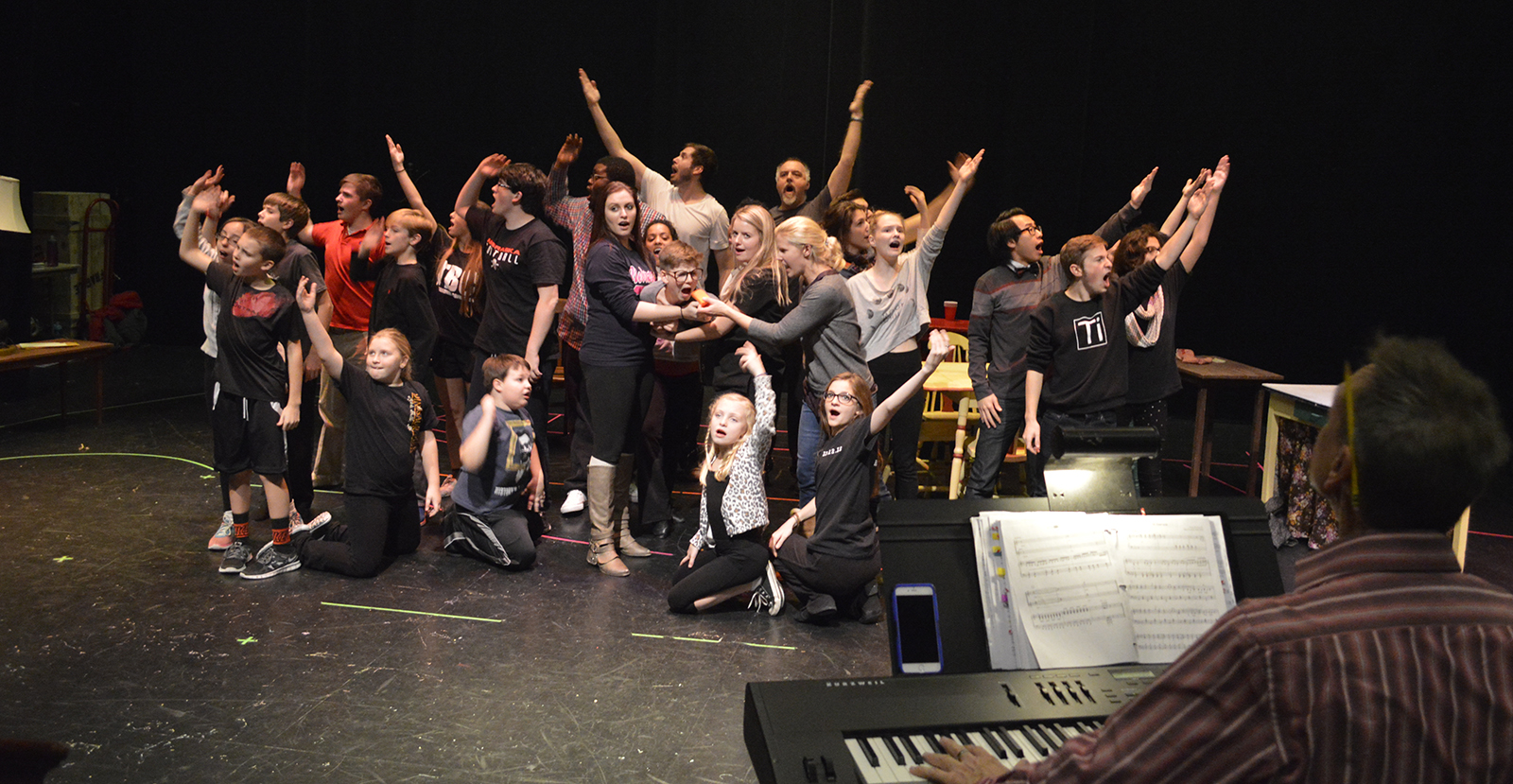 A group of students pose dramatically with their arms up as they sing in a rehearsal space while a music director at the piano watches them in the foreground.