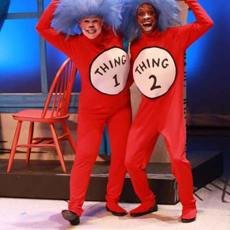 Two actors raise their fists in the air on stage while costumes in blue wigs and red onesies that read 