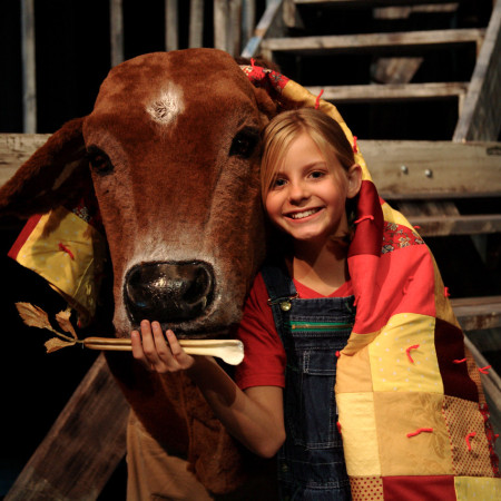 A youth actor holds a celery prop up to a large cow puppet face while both she and the cow have a quilt draped over them.