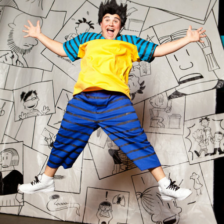A wigged actor playing Big Nate jumps high into the air with his hands and legs out wide. Backdrop on stage is black and white illustrations from the Big Nate book series.
