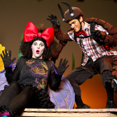 An actor dressed as a bug scares an actor dressed as a fly on stage. The fly has a large red bow, red lips, and purple wings.
