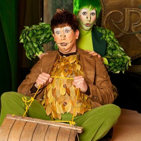Actors portraying Frog & Toad sit on a sled on stage. Both actors have worried expressions on their faces and foliage on their costumes.