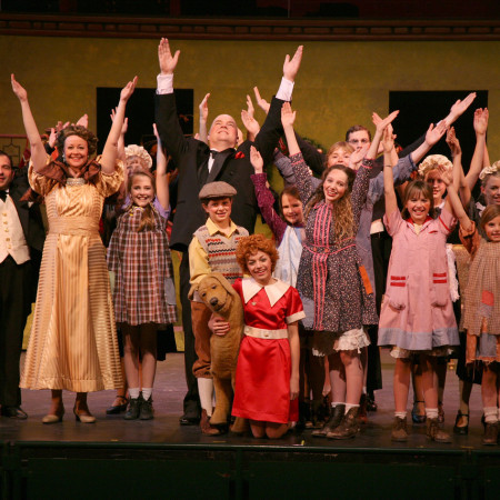 The cast of Annie all hold their arms overhead while surrounding Annie and her dog on stage.