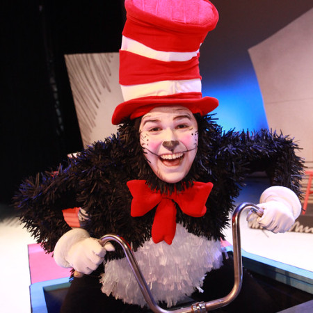 An actor portraying the Cat in the Hat leans over the handlebars of a bicycle on stage.