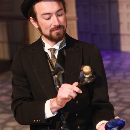 Actor portrays Sherlock Holmes in a suit and waistcoat while looking through a magnifying glass.