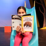 Nivi Varanasi as Vita in HOW TO BUILD AN ARK. PHOTO DESCRIPTION: A youth actor wearing a pink shirt, plaid vest, pink leggings, and yellow high-top tennis shoes sits on a turquoise playground slide while reading a National Geographic magazine. The cover image shows an armadillo with a headline about the Photo Ark.