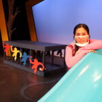 Nivi Varanasi as Vita in HOW TO BUILD AN ARK. PHOTO DESCRIPTION: A youth actor wearing a pink shirt leans alongside a turquoise playground slide. A black platform decorated with red, yellow and blue monkey figures can be seen behind her.