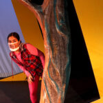 Nivi Varanasi as Vita in HOW TO BUILD AN ARK. PHOTO DESCRIPTION: A youth actor wearing a plaid vest, pink shirt, and pink leggings sits peeks out from behind a large yellow set piece reminiscent of the National Geographic magazine border.