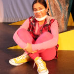 Nivi Varanasi as Vita in HOW TO BUILD AN ARK. PHOTO DESCRIPTION: A youth actor wearing a plaid vest, pink shirt, pink leggings, and yellow high-top tennis shoes sits cross-legged beside a large yellow set piece reminiscent of the National Geographic magazine border.