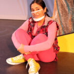 Nivi Varanasi as Vita in HOW TO BUILD AN ARK. PHOTO DESCRIPTION: A youth actor wearing a plaid vest, pink shirt, pink leggings, and yellow high-top tennis shoes sits cross-legged beside a large yellow set piece reminiscent of the National Geographic magazine border.
