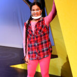 Nivi Varanasi as Vita in HOW TO BUILD AN ARK. PHOTO DESCRIPTION: A youth actor wearing a plaid vest, pink shirt, and pink leggings stands beside a large yellow set piece reminiscent of the National Geographic magazine border.