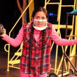 Nivi Varanasi as Vita in HOW TO BUILD AN ARK. PHOTO DESCRIPTION: A youth actor wearing a plaid vest and pink shirt, holding a large camera. She is shrugging in a gesture that says 