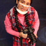 Nivi Varanasi as Vita in HOW TO BUILD AN ARK. PHOTO DESCRIPTION: A youth actor wearing a plaid vest, pink shirt and pink looks up. She is holding a large camera.