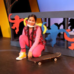 Nivi Varanasi as Vita in HOW TO BUILD AN ARK. PHOTO DESCRIPTION: A youth actor wearing a plaid vest, pink shirt and pink kneels by a skateboard. In the background can be seen a platform decorated with brightly-colored cutouts of monkey figures.