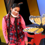Nivi Varanasi as Vita in HOW TO BUILD AN ARK. PHOTO DESCRIPTION: A youth actor wearing a plaid vest, pink shirt and pink leggings looks at an orange and black Monarch butterfly puppet. In the background can be seen a tree and a platform decorated with brightly-colored cutouts of monkey figures.