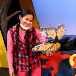 Nivi Varanasi as Vita and Tamia Renée as the Monarch Butterfly in HOW TO BUILD AN ARK. PHOTO DESCRIPTION: A youth actor wearing a plaid vest, pink shirt and pink leggings looks at an orange and black Monarch butterfly puppet that has landed on the lens of her camera. The butterfly puppeteer wears all black clothing and an orange mask. In the background can be seen a tree and a platform decorated with brightly-colored cutouts of monkey figures.