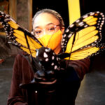 Tamia Renée as the Monarch butterfly in HOW TO BUILD AN ARK. PHOTO DESCRIPTION: A female actor wearing a black costume and orange face mask holds out an orange and black Monarch butterfly puppet.
