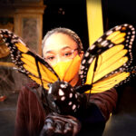 Tamia Renée as the Monarch butterfly in HOW TO BUILD AN ARK. PHOTO DESCRIPTION: A female actor wearing a black costume and orange face mask holds out an orange and black Monarch butterfly puppet.