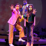 Photo description: Three actors strike a dramatic pose with their arms outstretched. A male actor wears a sparkly pink blazer over a gold vest and pants with a pink button-up shirt. Another male actor is wearing a blue military-inspired jacket over a yellow and blue jumpsuit with bright blue boots and a blue mask. A female actor is wearing a greet jacket embellished with pink sequins and long pink fringe along the sleeves and back of the jacket. She wears striped grey pants with pink sequin trim and pink high-heeled boots.