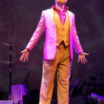 Photo description: A male actor wears a sparkly pink blazer over a gold vest and pants with a pink button-up shirt. He is standing on a step singing earnestly, looking up to the balcony with his arms outstretched near his hips.