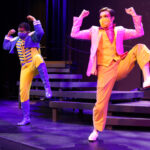 Photo description: Two male actors dance in front of a set of stairs. Their arms are by their heads, bent into a flexed 
