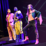 Photo description: Three actors strike a dramatic pose with their arms outstretched and their hands in fists. A male actor wearing a sparkly pink blazer over a gold vest and pants with a pink button-up shirt has his arm stretched to the left. Another male actor is wearing a blue military-inspired jacket over a yellow and blue jumpsuit with bright blue boots and a blue mask; his arm is stretched out to the camera. A female actor is wearing a greet jacket embellished with pink sequins and long pink fringe along the sleeves and back of the jacket. She wears striped grey pants with pink sequin trim and pink high-heeled boots.