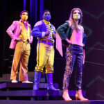 Photo description: Three actors stand on a set of stairs with their hands on their hips. A male actor wears a sparkly pink blazer over a gold vest and pants with a pink button-up shirt. Another male actor is wearing a blue military-inspired jacket over a yellow and blue jumpsuit with bright blue boots and a blue mask. A female actor is wearing a greet jacket embellished with pink sequins and long pink fringe along the sleeves and back of the jacket. She wears striped grey pants with pink sequin trim and pink high-heeled boots.