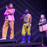 Photo description: Three actors strike a dramatic pose with their arms outstretched. A male actor wears a sparkly pink blazer over a gold vest and pants with a pink button-up shirt. Another male actor is wearing a blue military-inspired jacket over a yellow and blue jumpsuit with bright blue boots and a blue mask. A female actor is wearing a greet jacket embellished with pink sequins and long pink fringe along the sleeves and back of the jacket. She wears striped grey pants with pink sequin trim and pink high-heeled boots.