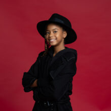 Young girl in black fedora and trench coat posing with her arms crossed.