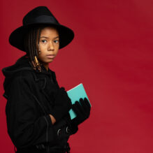 Young girl with black fedor and trench coat pulls a light blue notebook from inside her jacket.