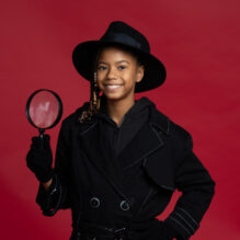 Young girl wearing a black fedora and trench coat holds a magnifying glass up.