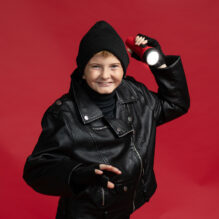 A young boy in a black stocking cap and leather jacket poses with a red flashlight by his head.