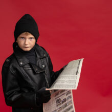 A young boy in a black stocking cap and leather jacket looks at the camera holding a sheet of newspaper.