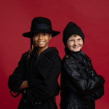 A young girl in a black fedora and trench coat stands back to back with a young boy with a black stocking cap and leather jacket.