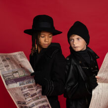 A young girl and boy wearing spy gear stand back to back holding sheets of newspaper.