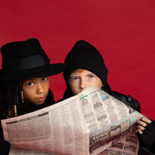 A young girl and boy in spy gear peek over the top of a newspaper.