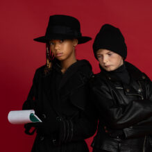 A young girl and boy in spy gear stand shoulder to shoulder. The girl is holding a notebook open.