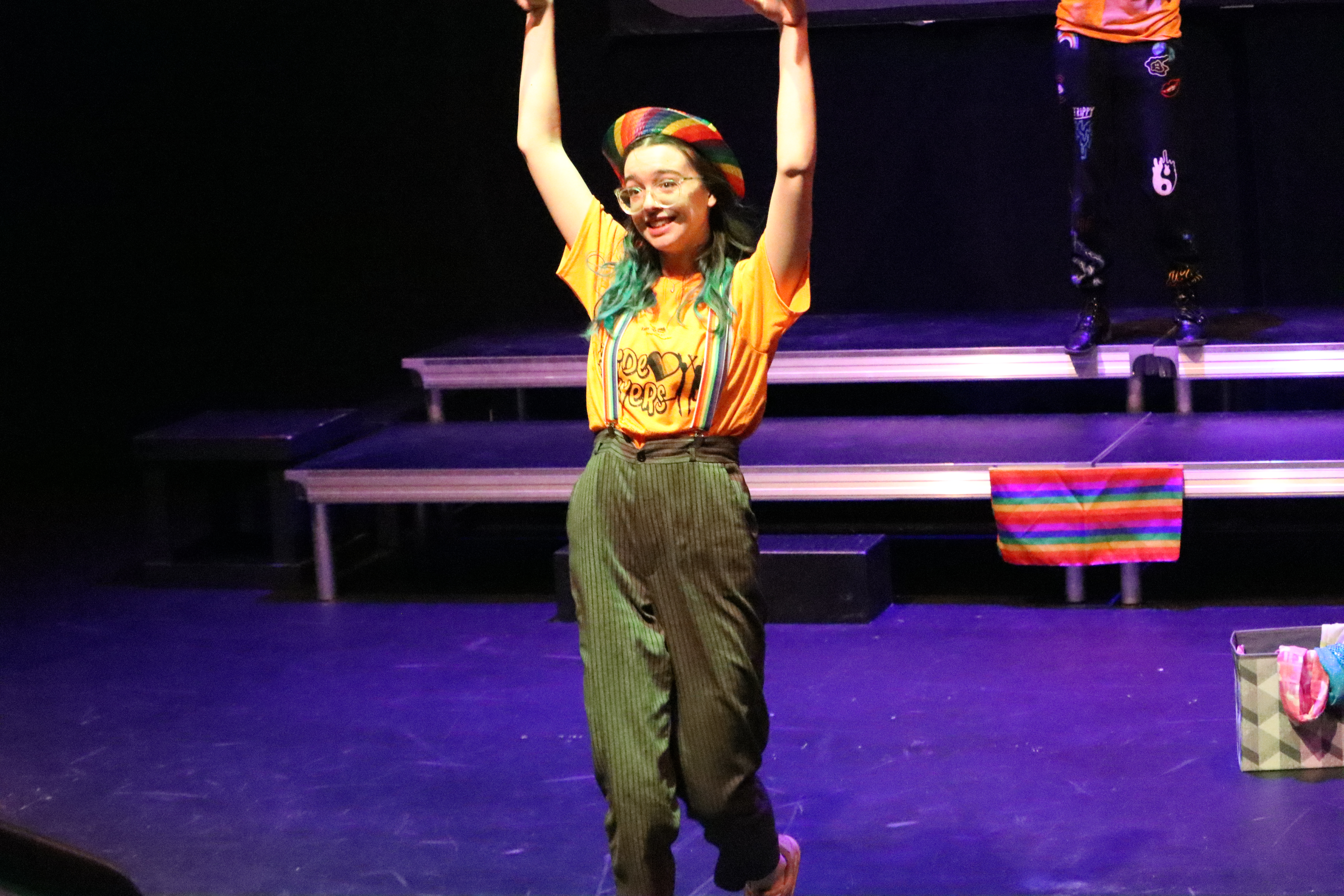 A Teens N Theater performer smiling while performing a routine. They are wearing a rainbow beret, a yellow t-shirt with green pants and suspender.