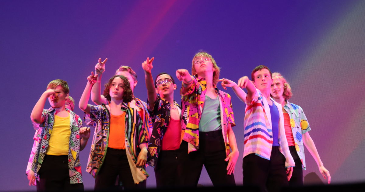 A group of costumed performers in formation singing a musical number. Their costumes are short-sleeve button down shirts.