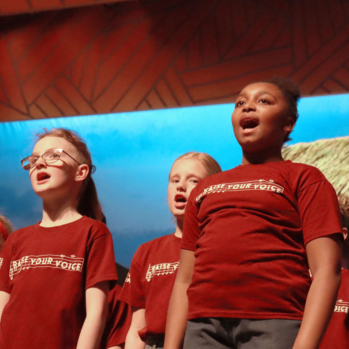 A diverse group of students performing a musical number wearing matching Raise Your Voice t-shirts.