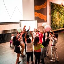 A group of people, including adults and children, stand in a circle raising their hands and large glowing balls in the air. They are engaged in an interactive experience in front of a screen displaying abstract line drawings. The environment has a modern aesthetic with a green wall and large windows letting in natural light, suggesting a public or cultural space.