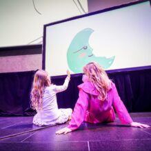 A young girl and an adult with long, wavy hair sit on the floor in front of a large screen displaying an animated green crescent moon with a face. The girl, wearing a light-colored dress, reaches out toward the screen with a small paddle, interacting with the projected image. The adult, dressed in a pink outfit, sits beside her, observing the scene.