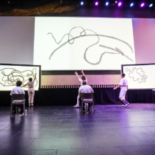 A live performance on a stage features five performers dressed in white, interacting with abstract line drawings projected on three large screens. Two performers are seated, facing the screens, while three are standing, gesturing or drawing on the screens. The stage is illuminated with purple and warm lighting, with a textured backdrop featuring wavy gold accents. The abstract drawings consist of thick black lines forming loops and curves.