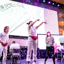 A live performance on a stage features five performers dressed in white, interacting with abstract line drawings projected on three large screens. The stage is illuminated with purple and warm lighting, with a textured backdrop featuring wavy gold accents. The abstract drawings consist of thick black lines forming loops and curves.