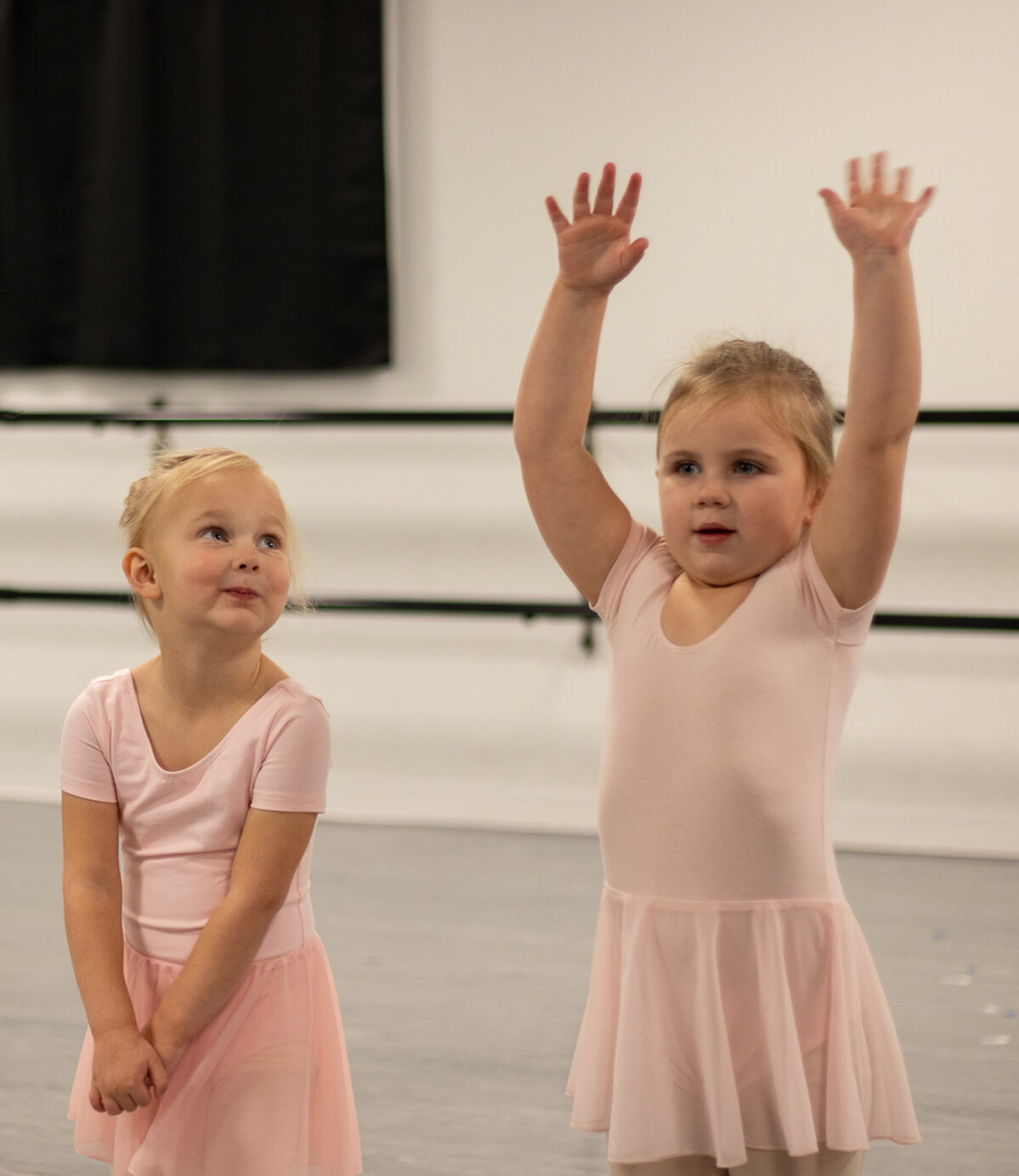 Two young girls in light pink leotards and skirts stand in a dance studio. The girl on the left clasps her hands shyly and looks up with a smile, while the girl on the right raises both arms high above her head, looking focused. The background shows a ballet barre and a white wall.